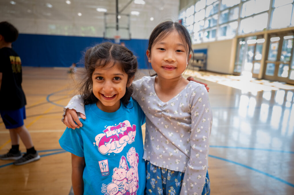 Two girls with their arms around eachother smiling at the camera inside a gym.