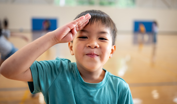 A boy smiles and salutes in front of the camera.